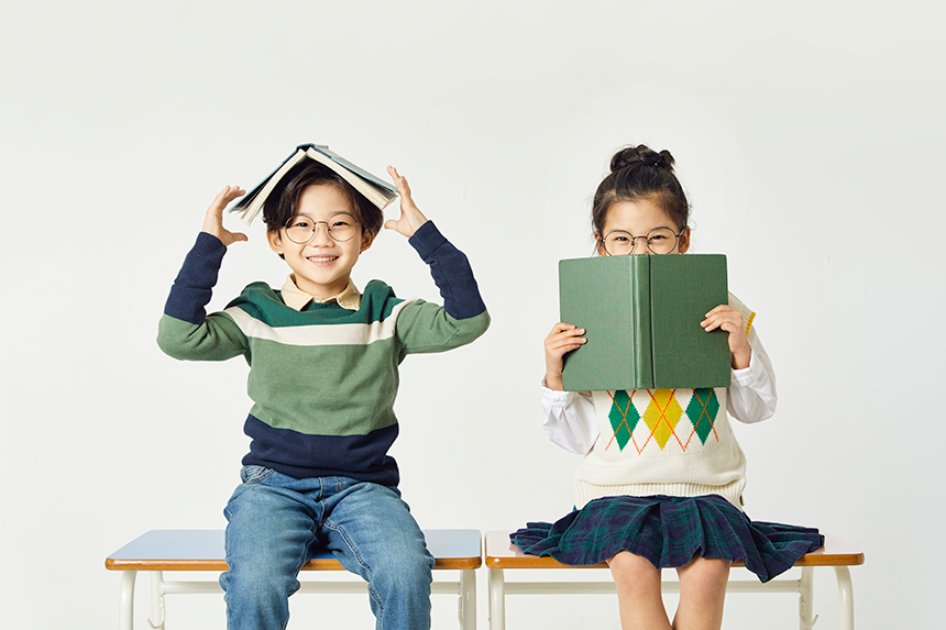 각자 책상위에 앉아, 책을 머리에 쓴 남자 어린이와 책으로 하관을 가린 여자 어린이가 활짝 웃고 있는 모습.